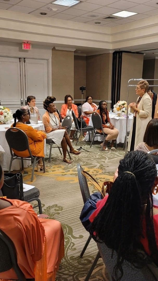 A woman speaks to an attentive group seated at round tables in a conference room.